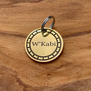 May include: A gold-colored oval dog tag with a black engraved border and the text "W'Kabi" in black.