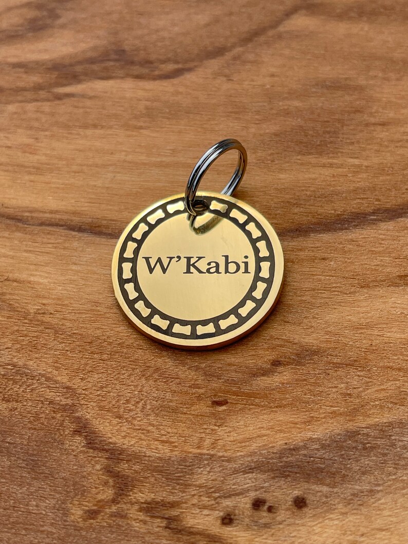 May include: A gold-colored oval dog tag with a black engraved border and the text "W'Kabi" in black.