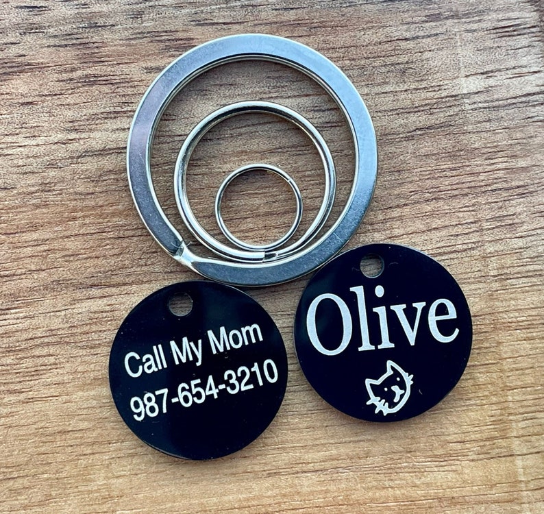 May include: Two black round pet ID tags with white text. One tag says "Call My Mom 987-654-3210" and the other tag says "Olive" with a small cat icon.
