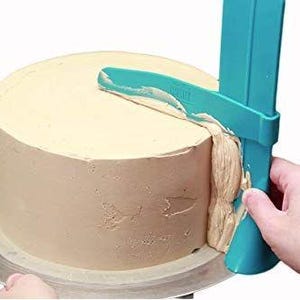 May include: A turquoise cake scraper tool is being used to smooth the edges of a frosted cake. The cake is on a silver cake turntable.