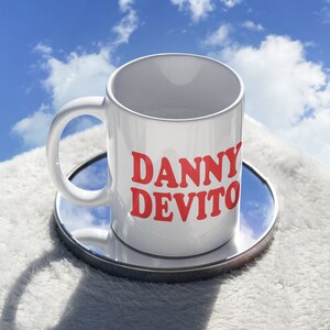 Puede incluir: Taza de cerámica blanca con el nombre "DANNY DEVITO" impreso en rojo, colocada sobre un pequeño posavasos redondo y espejado. La taza tiene un asa curvada y está sobre una superficie blanca y esponjosa, con un cielo azul y nubes al fondo.