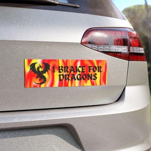 May include: A rectangular bumper sticker with a fiery background of yellow, orange, and red. A black dragon silhouette is on the left, and the text "I BRAKE FOR DRAGONS" is on the right. The sticker is affixed to the back of a silver car.