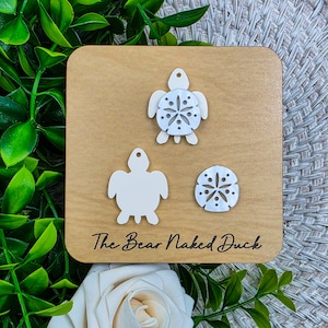 May include: A wooden square with three white sea-themed cutouts: two turtles and a sand dollar. The text "The Bear Naked Duck" is printed in black. The background includes green leaves and a white flower.