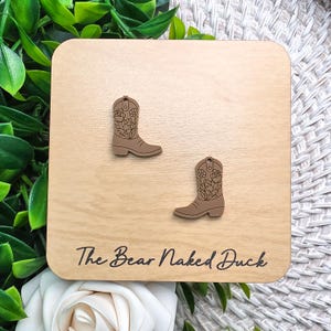 May include: A pair of light brown cowboy boot earrings featuring a delicate black floral line art design. The earrings are displayed on a square wooden card with the text The Bear Naked Duck printed in black script at the bottom.