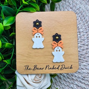 May include: A pair of dangle earrings featuring a white ghost shape with a speckled texture, topped with an orange bow and a black flower with a gold center. The earrings are displayed on a square wooden card labeled The Bear Naked Duck.