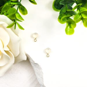 May include: Two pairs of small, delicate gold-colored earrings with a round, translucent stone. The earrings are set against a white background, with green foliage and a white rose in the corners.
