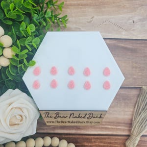 May include: Ten pink Easter egg-shaped resin embellishments with a textured design. The embellishments are arranged in three rows on a white hexagonal surface.