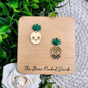 May include: Gold skull-shaped earrings with a green pineapple top, displayed on a wooden plaque. The earrings have a lattice design and are set against a backdrop of green foliage and a white rose. The text "The Bear Naked Duck" is visible.