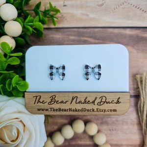 May include: A pair of black and white checkered bow earrings on a white background. The earrings are displayed on a wooden stand with the text "The Bear Naked Duck" and the website address "www.TheBearNakedDuck.Etsy.com".