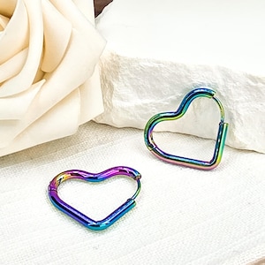 May include: Two heart-shaped hoop earrings with a rainbow iridescent finish. The earrings are made of metal and have a hinged closure. The earrings are displayed on a white surface.