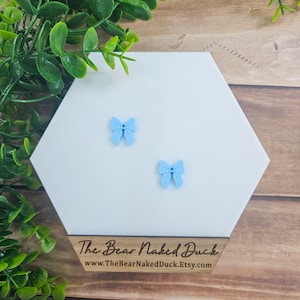 May include: Two light blue bow-shaped wooden earrings on a white hexagonal display card. The card has the text "The Bear Naked Duck" and "www.TheBearNakedDuck.Etsy.com" printed on it.