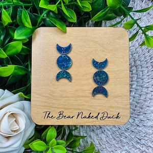 May include: Two pairs of blue moon phase earrings on a wooden display. The earrings have a shimmering, iridescent finish with star-like details. The wooden display has the text "The Bear Naked Duck" in black script.