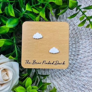 May include: A pair of cloud-shaped earrings with a glittery white finish, displayed on a wooden square. The text "The Bear Naked Duck" is printed in black script at the bottom of the wooden square.