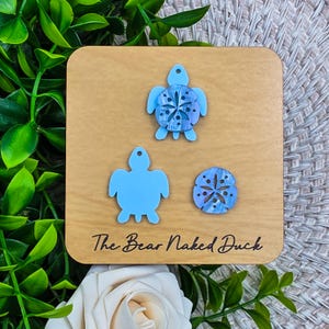 May include: A wooden square with three sea-themed acrylic shapes: two blue turtles and a blue and purple sand dollar. The text "The Bear Naked Duck" is printed in black on the wood. The shapes are for earrings or other jewelry.