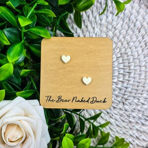 May include: A pair of heart-shaped earrings in a light cream color are displayed on a small, square, light brown card. The card has the text "The Bear Naked Duck" printed in black. The earrings are set against a backdrop of green foliage and a white rose.