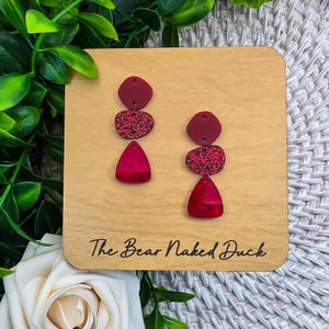 May include: Earrings displayed on a wooden plaque. The earrings are a deep red color, with a mix of solid shapes and glittery textures. The plaque has the text "The Bear Naked Duck" in a handwritten style.