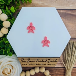 May include: Two pink turtle charms on a white background. The turtles are made of a delicate, intricate design. The background is a white hexagon shape. The text "The Bear Naked Duck" and a website address are visible at the bottom of the image.
