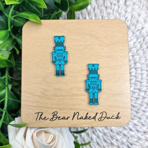May include: A pair of teal and blue acrylic nutcracker earrings. The earrings are displayed on a light brown wooden square with the text "The Bear Naked Duck" in black script. The earrings are a festive accessory.