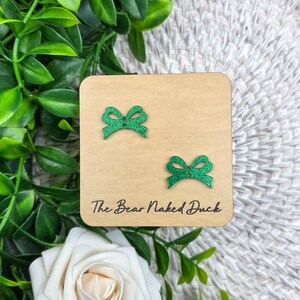 May include: A pair of green glitter bow earrings on a light brown wooden square. The earrings are small, with a bow design. The text "The Bear Naked Duck" is printed on the wooden square.