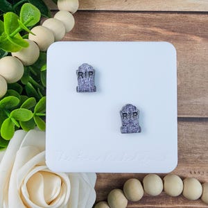 May include: A pair of small, gray tombstone-shaped earrings with the letters "RIP" engraved on them. The earrings are displayed on a white square surface, with a wooden background and decorative elements.