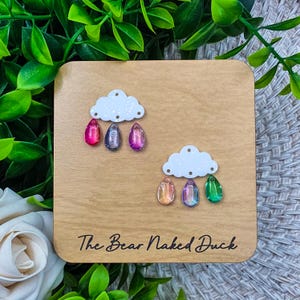 May include: Wooden square with two white cloud cutouts, each with three colorful teardrop-shaped embellishments. The text "The Bear Naked Duck" is printed in black script at the bottom of the wood.