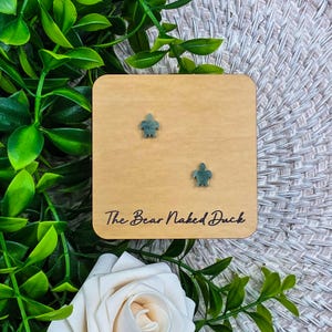 May include: A pair of small, teal turtle-shaped earrings displayed on a light brown wooden square. The text "The Bear Naked Duck" is printed on the wood. The earrings are set against a backdrop of green leaves and a white rose.
