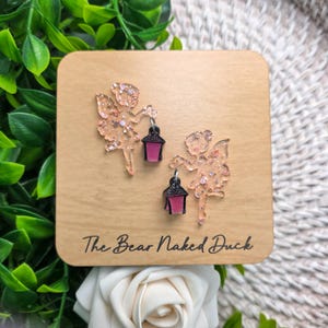 May include: A pair of glittery, translucent peach-colored fairy earrings, each holding a small black lantern with a pink center. The earrings are displayed on a square tan card with the text The Bear Naked Duck printed in black script.