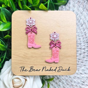 May include: A pair of dangle earrings featuring a pink cowboy boot with a floral line art design, topped with a dark pink bow and a glittery flower with a central pearl bead. The earrings are displayed on a tan card labeled The Bear Naked Duck.