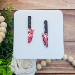 May include: A pair of novelty earrings shaped like small knives. The blades are red with a splatter pattern, and the handles are black. The earrings are displayed on a white surface.