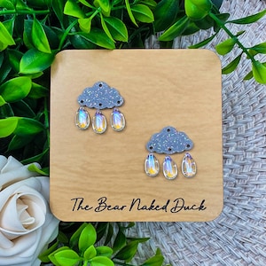 May include: A pair of cloud-shaped earrings with iridescent raindrop charms. The earrings are displayed on a wooden square with the text "The Bear Naked Duck" in a cursive font.