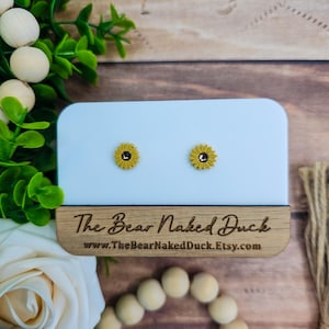May include: A pair of yellow sunflower stud earrings with a black center, displayed on a light blue card. The earrings are on a wooden base with the text "The Bear Naked Duck" and a website address.