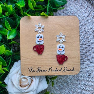 May include: A pair of dangle earrings featuring a silver-toned snowflake charm, a white snowman head with an orange nose, and a red glittery coffee mug charm. The earrings are displayed on a light wood square card labeled The Bear Naked Duck.