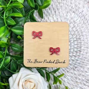 May include: A pair of red glitter bow earrings on a light brown wooden display. The earrings are small and detailed, with a classic bow shape. The text "The Bear Naked Duck" is printed on the display.