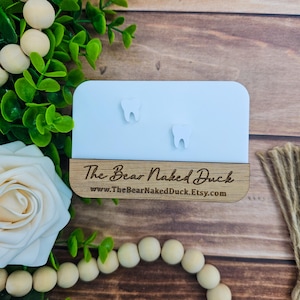 May include: White tooth-shaped earrings on a white display card with a wooden base. The base has the text "The Bear Naked Duck" and the website address "www.TheBearNakedDuck.Etsy.com".