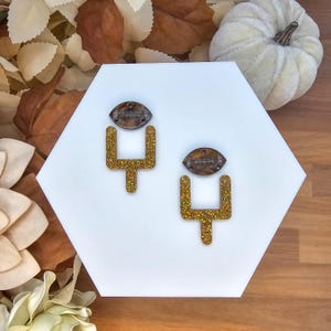 May include: A pair of gold glitter football-shaped earrings with brown tops. The earrings are displayed on a white hexagonal surface, with a small white pumpkin and autumn leaves in the background. The earrings are a fun accessory.