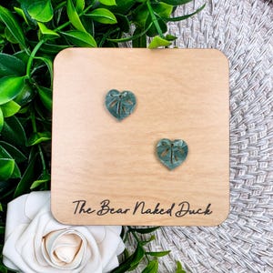 May include: Two teal heart-shaped earrings with a dragonfly design are displayed on a light wooden square. The earrings are set against a backdrop of green foliage and a white rose. The text "The Bear Naked Duck" is printed on the wood.