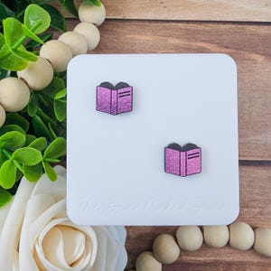 May include: Two pink glitter book-shaped earrings on a white surface. The earrings have black spines and two lines on the pages. The background includes wooden beads, greenery, and a white flower.