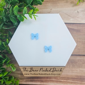 May include: A pair of light blue bow earrings on a white hexagonal display stand. The stand is on a wooden surface. The text "The Bear Naked Duck" and "www.TheBearNakedDuck.Etsy.com" is printed on the stand.