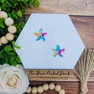 May include: Two colorful starfish-shaped earrings with a vibrant mix of blue, yellow, and pink hues. They are displayed on a white hexagonal surface with a wooden base that has the text "The Bear Naked Duck" and a website address.