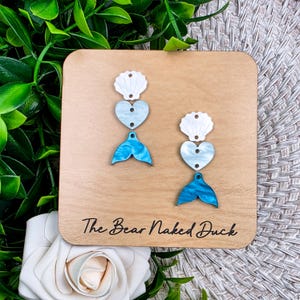 May include: A pair of mermaid-themed earrings displayed on a wooden square. Each earring features a white seashell, a light blue heart, and a blue mermaid tail. The wooden square has the text "The Bear Naked Duck" in black.