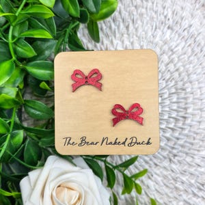 May include: A pair of red glitter bow earrings on a light brown wooden square. The earrings are small and detailed, with a textured, sparkly finish. The text "The Bear Naked Duck" is printed on the wood.