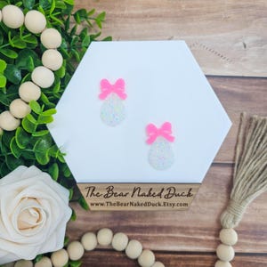 May include: A pair of white glitter earrings with pink bows. The earrings are on a white hexagonal display stand. The text "The Bear Naked Duck" and a website address are visible on the stand.