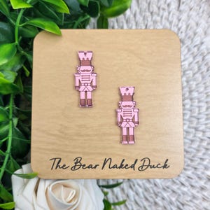 May include: Pink nutcracker earrings on a wooden display card. The earrings are shaped like toy soldiers with detailed features. The card has the text "The Bear Naked Duck" in a cursive font.