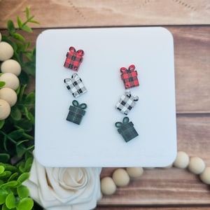 May include: Six small gift-shaped earrings in red, black and white, and green plaid patterns are arranged on a white square. The earrings are on a white surface, with greenery and wooden beads in the background.