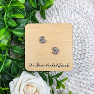 May include: A pair of gray and black spotted crescent-shaped stud earrings on a wooden display card. The card has the text "The Bear Naked Duck" printed in black. The background includes green leaves and a white rose.