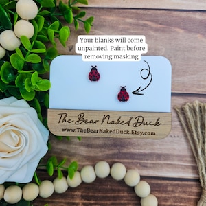 May include: Two ladybug-shaped earring blanks with red and black details are displayed on a white card. The card is mounted on a wooden base with the text "The Bear Naked Duck" and website address. Text above the earrings reads "Your blanks will come unpainted."