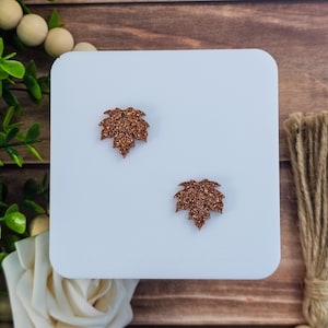 May include: Two glittery copper-colored maple leaf-shaped embellishments on a white square surface. The leaves are detailed with multiple points and a textured, sparkling finish. The background is a wooden surface with decorative elements.