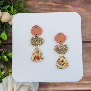 May include: A pair of handmade earrings with a layered design. The earrings feature a peach-colored top piece, a gold glitter middle piece, and a floral patterned bottom piece. The floral pattern includes orange, yellow, and white flowers. The earrings are displayed on a white surface.