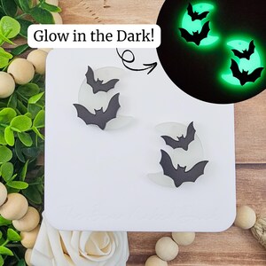 May include: A pair of bat-shaped earrings with a crescent moon design. The earrings are black and white and glow in the dark. The image includes the text "Glow in the Dark!"