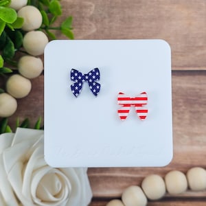 May include: Two small bow-shaped accessories. One is navy blue with white stars, and the other is red with white stripes. The bows are on a white surface with the text "The Bear Naked Duck" at the bottom.